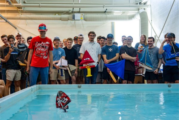 Caleb Ulman, in red shirt at left, watches his sailboat travel across a shallow pool, large crowd behind him