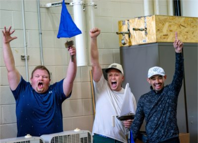 three engineering students cheer with arms in the air, one on left is holding a model sailboat