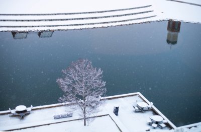 A snowy scene, seen from above. Looking down on snow covered tables, a tree, and a pond. The top of a building and carillon tower reflect on the pond.