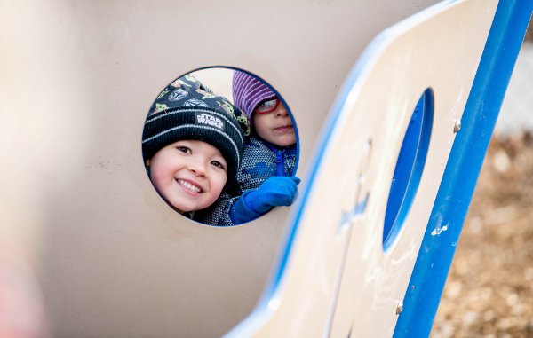 Two children look through a round opening of a piece of playground equipment at the camera. 
