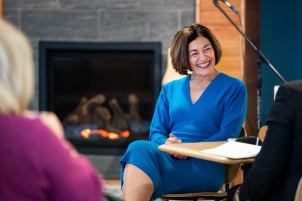 A woman dressed in blue laughs. There is a fireplace glowing behind her and people with their backs to the camera in front of her.  