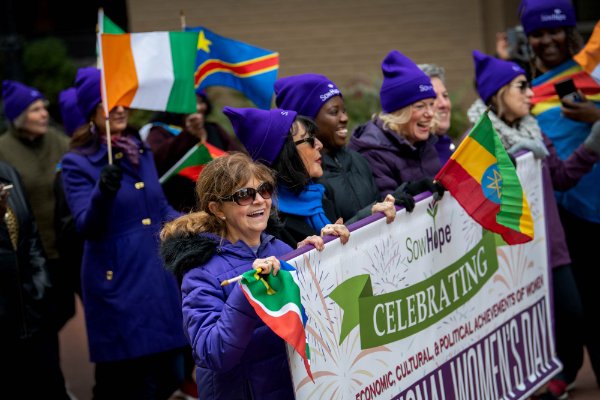 A group of women march wearing purple hats holding a banner and colorful international flags during a march through town.