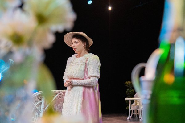 A actor wearing a white and pink dress and straw hat performs on stage near white wicker furniture during a play.