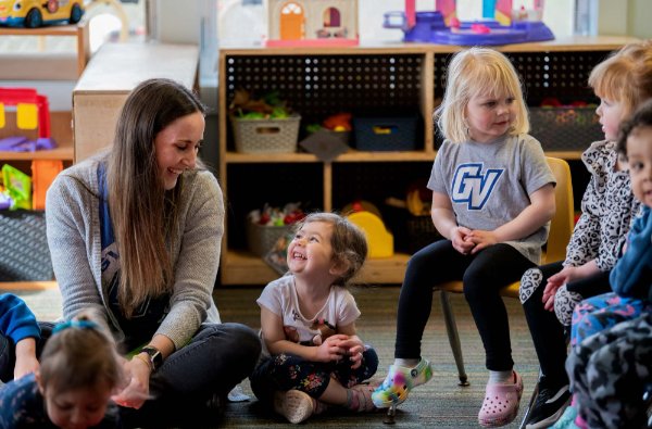 A preschool teacher and a young child smile at each other while sitting in a circle among other young children in a preschool classroom.