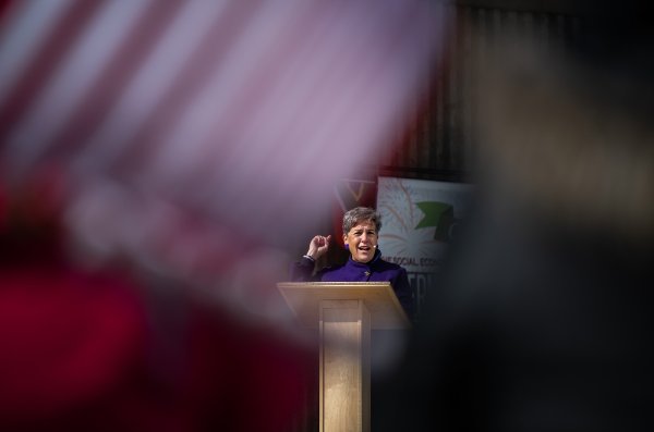 A speaker framed by the American Flag stands behind a lectern and speaks to a crowd.