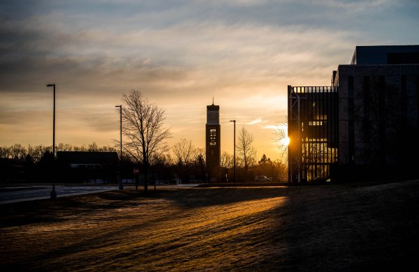 The sunrise illuminates a carillon tower and library building on a college campus.