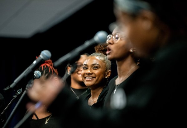 A person, seen through microphones and other people singing, smiles toward the camera during a choir performance.