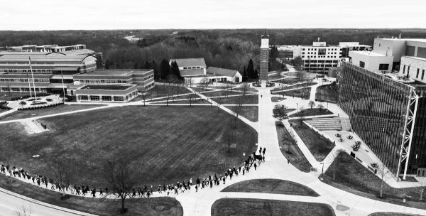 Photo from a drone of a line of people walking on a sidewalk curving toward a clock tower.