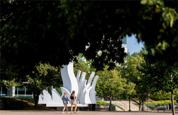 Two people walk past a white sculpture framed by green trees on a college campus. 