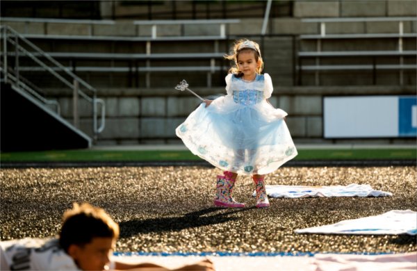  A little child wearing a princess dress, rubber boots, crown and wand twirls around on a football field. 