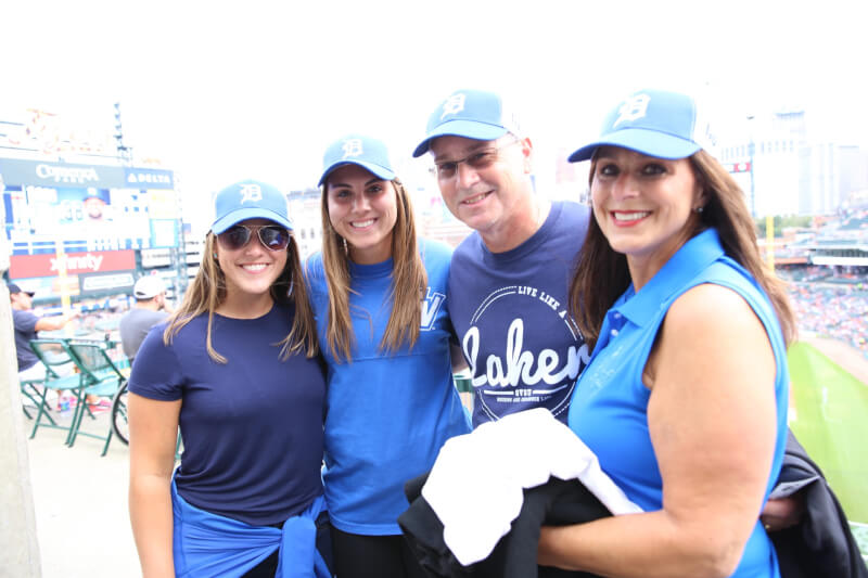  Four GVSU Alumni standing, smiling with the baseball field in the background