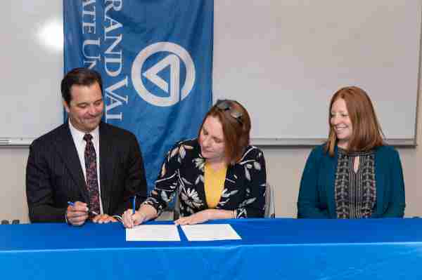 Two people are seated at a table with a blue table cloth. One person signs paper while another looks on. A Grand Valley State University flag hangs behind them.