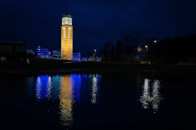 Cook Carillon Tower lit up with trees lit with blue lights surrounding it; tower is reflected in Zumberge pond.