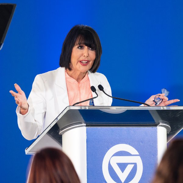 A person standing at a podium with the GVSU logo gestures with both hands while speaking. The person is holding eyeglasses in the left hand.