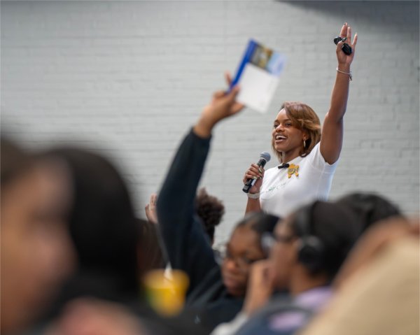 Sharnell James raises her left arm while giving a presentation, people seated at tables in front