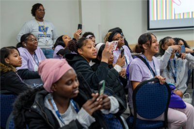 young girls seated, many taking phone photos, woman in back standing