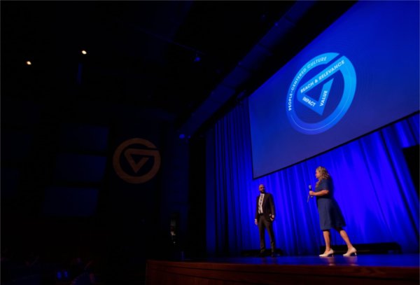 two people on stage, blue curtain and circle GV on projection screen