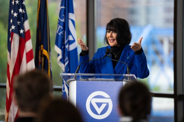 The president of GVSU addresses a crowd at a podium with flags and downtown Grand Rapids behind her. 
