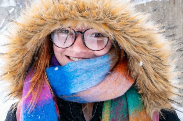 A college student wearing glasses and bundled up with a furry hood and colorful scarf braves the cold weather with a smile. 