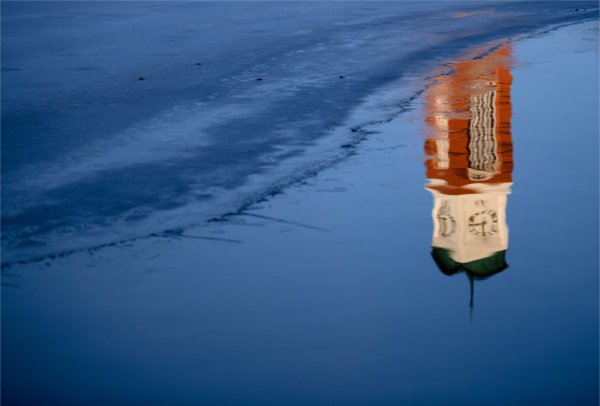  A carillon tower is reflected into blue water near a frozen section of a pond. 