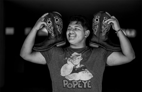 A portrait of a college student wearing a Popeye shirt holding cowboy boots on his shoulders. 