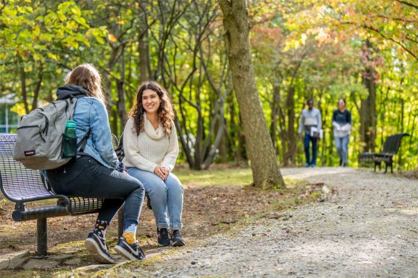 College students smile while sitting on a bench in a wooded area with other students walking in the background.  