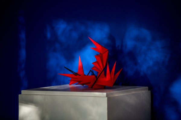  A red piece of art made up of spikes sits on a white square column against a dark blue background with shadows across the back. 
