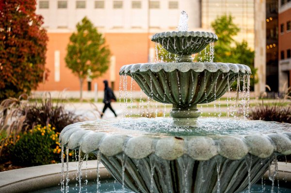 Water flows from a tiered fountain in the foreground while a person walks in front of a building in the background. 