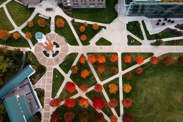  Drone angle of campus showing buildings, clock tower, orange and red trees and zig-zagging sidewalks. 