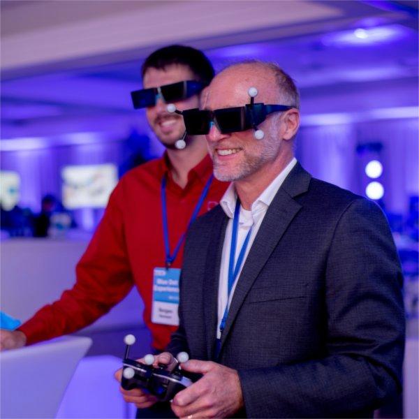 A man in a suit and another man in a red shirt wear augmented-reality glasses with tracking markers during a technology demonstration.