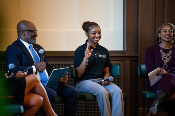 A person creates an "anchor" sign while people on either side smile. All are seated. The person to the left is holding a microphone.