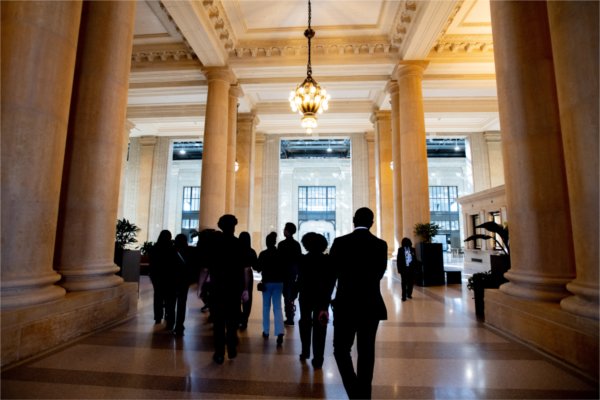 Several people are seen from behind as they walk through a room with tall pillars and intricate overhead lights.