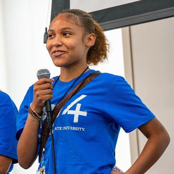 A young woman in a REP4 shirt holds a microphone and speaks as her team pitches their REP4 idea.