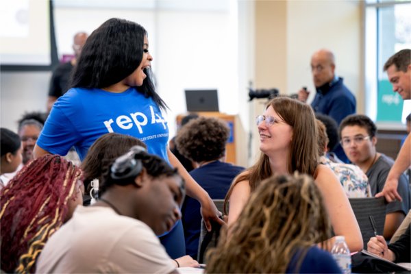 A GVSU student in a REP4 shirt chats with a table of high school students.