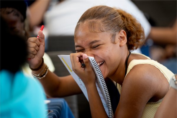 A young woman holds a notebook to her face as she laughs during a group activity.