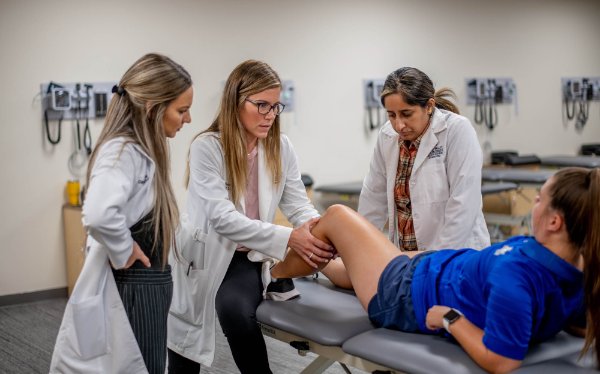 three nursing students gather around a table where a student in a blue shirt in laying; they are evaluating her left knee during a simulated exercise
