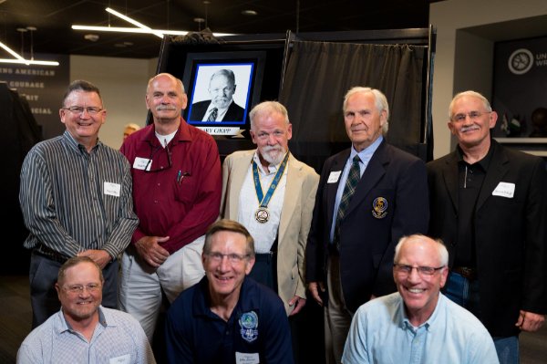 group pictured, five men standing, three men kneeling by a TV screen of Gary Chopp during the National Wrestling Hall of Fame induction ceremony