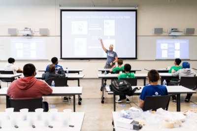 instructor leads a class of campers seated in chairs; everyone wearing masks