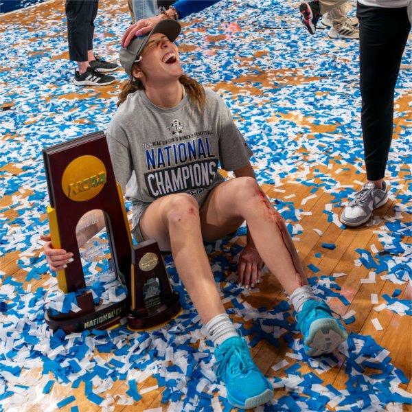 A smiling women's basketball player sits on a confetti-covered floor with her arm around the national championship trophy.