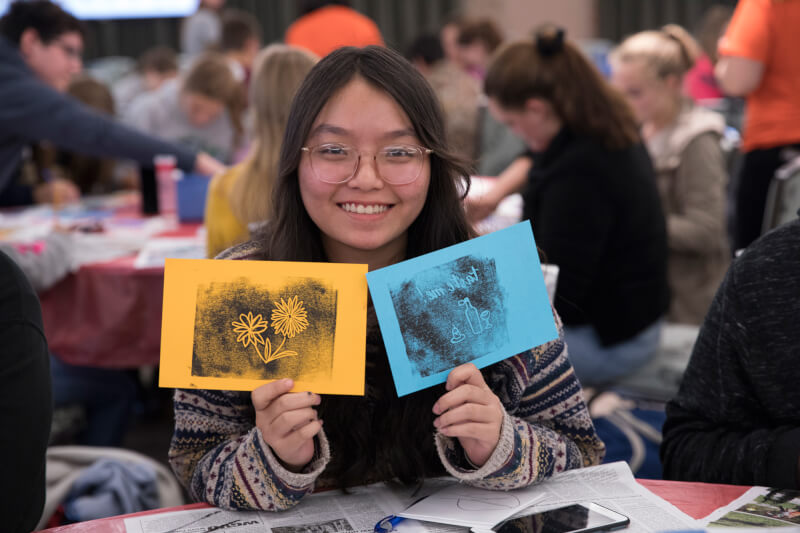 A student holds up two ink prints, one of a flower and one of a vase, after completing them in a hands-on workshop.