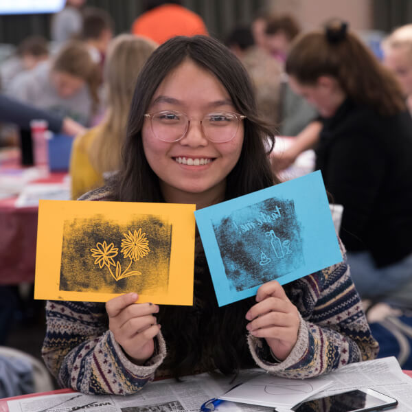 A student holds up two ink prints, one of a flower and one of a vase, after completing them in a hands-on workshop.