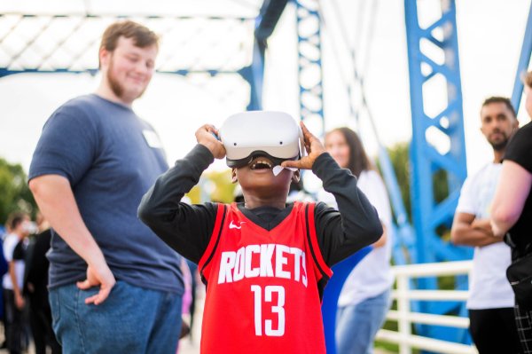 A person wearing a jersey that says "Rockets" and the number 13 looks through a VR headset. A person smiles in the background.