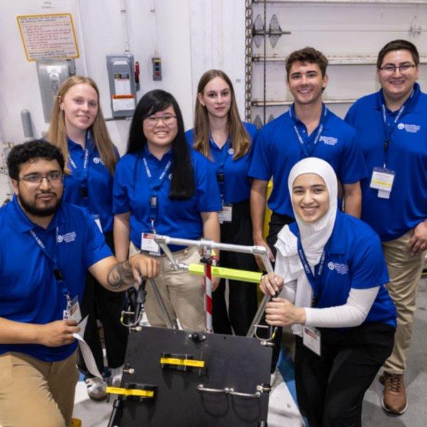 students in blue shirts stand and kneel by a machine they built