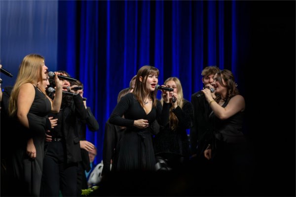 Student singers in black stand on stage performing with microphones