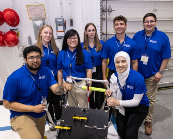 students in blue shirts stand and kneel by a machine they built