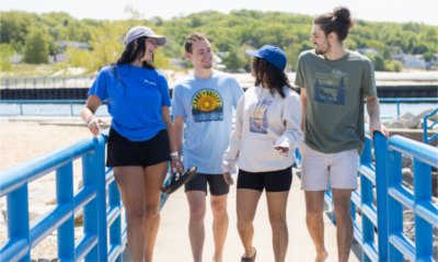 Four students wearing new Lake + Valley clothing items walk along the beach, laughing and talking together.