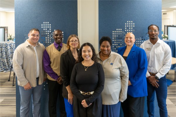 seven people standing in V formation in front of blue accent wall