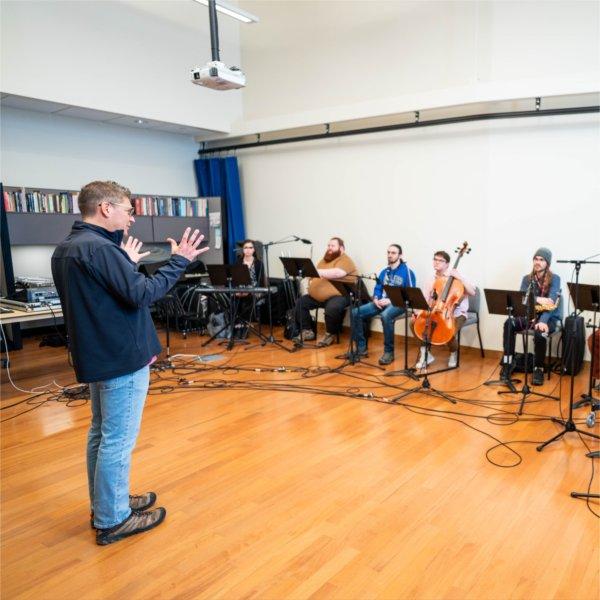 A person gestures with hands while standing before people seated with musical instruments in front of music stands.