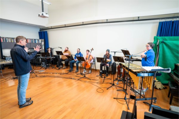 A person gestures while standing before people seated with musical instruments. Music stands are in front of them.