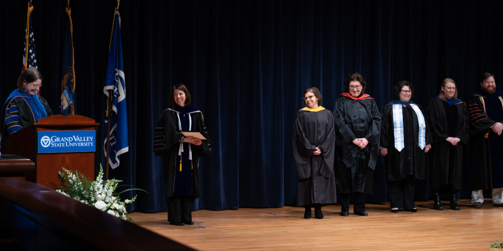 woman at podium with other faculty members in regalia on stage